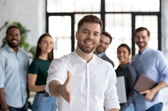 Hombre con camisa blanca extendiendo la mano, con un grupo de personas sonriendo detrás.